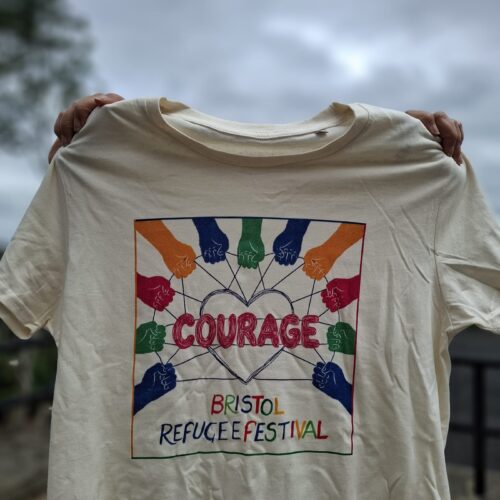 Cream Bristol Refugee Festival 2026 Courage T-shirt held up to show the colourful hands and heart design.